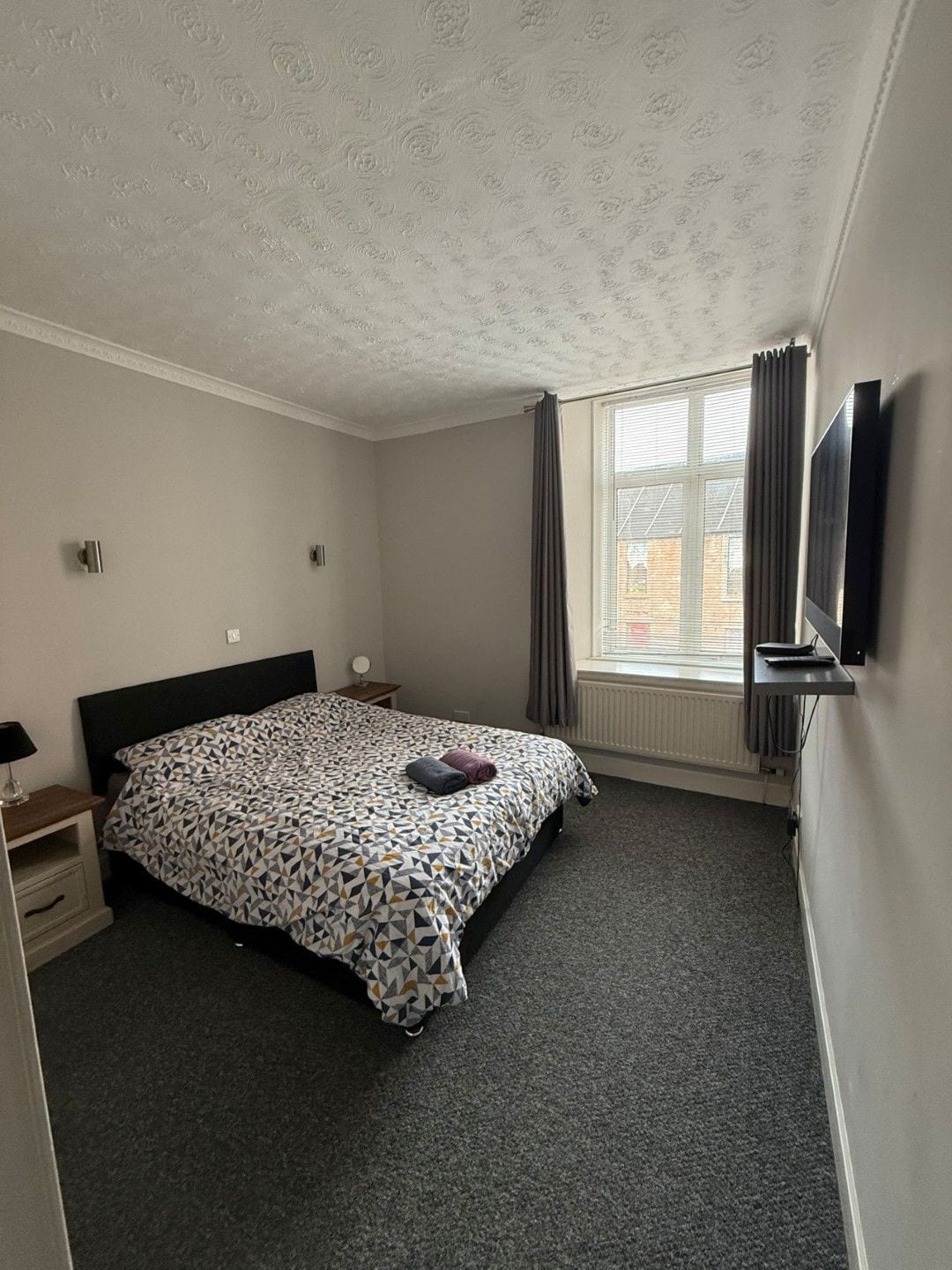 Een slaapkamer met een bed, nachtkastje, lamp, raam met gordijnen en een aan de muur bevestigde televisie.