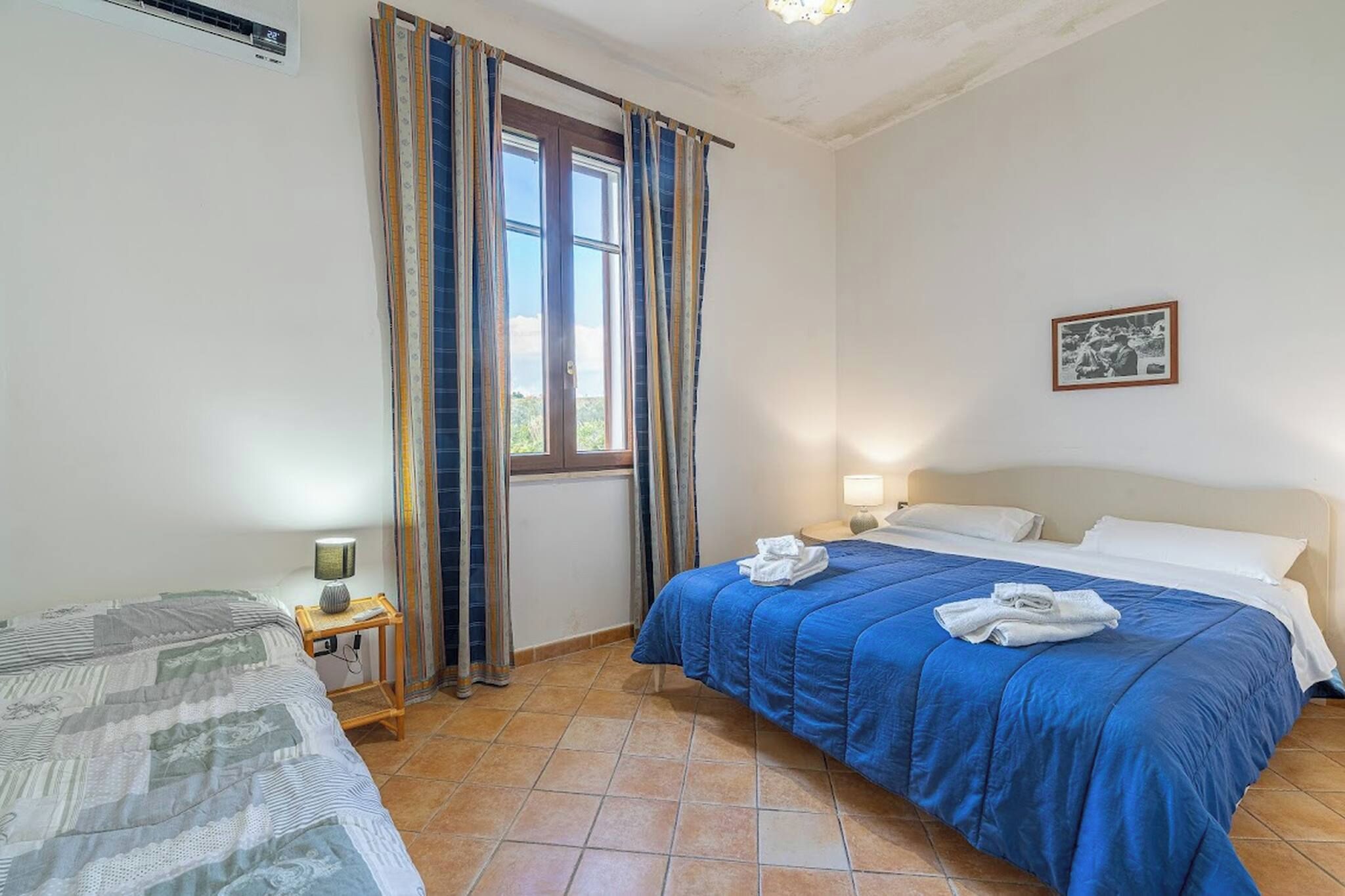 A bedroom with a bed, a bedside table with a lamp, a window with curtains, and a wall-mounted picture.