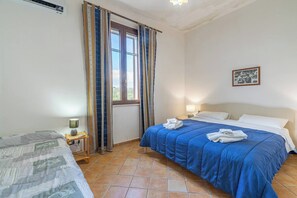 Un dormitorio con cama, mesita de noche con lámpara, ventana con cortinas e imagen colgada en la pared.