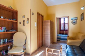 Ein Wohnzimmer mit einer Couch, einem Sessel, einem kleinen Tisch, einem Bücherregal, einer Tür und einem Fenster mit Vorhängen.