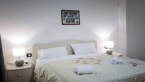 A neatly made bed with white linens and a floral-patterned duvet, two bedside lamps, a small round table with a lamp, and a framed picture on the wall. - Charming Villa with Lemon Garden Near Lido di Noto Beach (Noto)