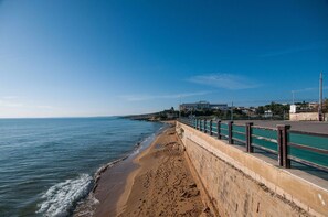 Uma passarela à beira-mar com barreira de concreto e corrimão de metal, que leva a uma praia arenosa.