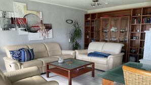 Sala de estar com sofás bege, mesa de centro de vidro, estante e abajur.
