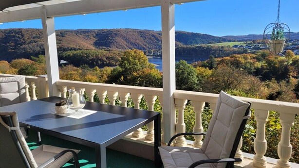 Área de estar coberta ao ar livre, com mesa e cadeiras, com vista para uma paisagem com colinas e um rio.