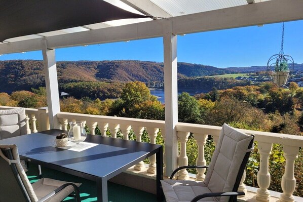 A covered outdoor seating area with a table and chairs, overlooking a scenic landscape with hills and a river. - Dream view of the Rursee (Simmerath-Woffelsbach)