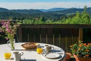 Mesa al aire libre con vista a la montaña, un jarrón de flores y un vaso de jugo de naranja.