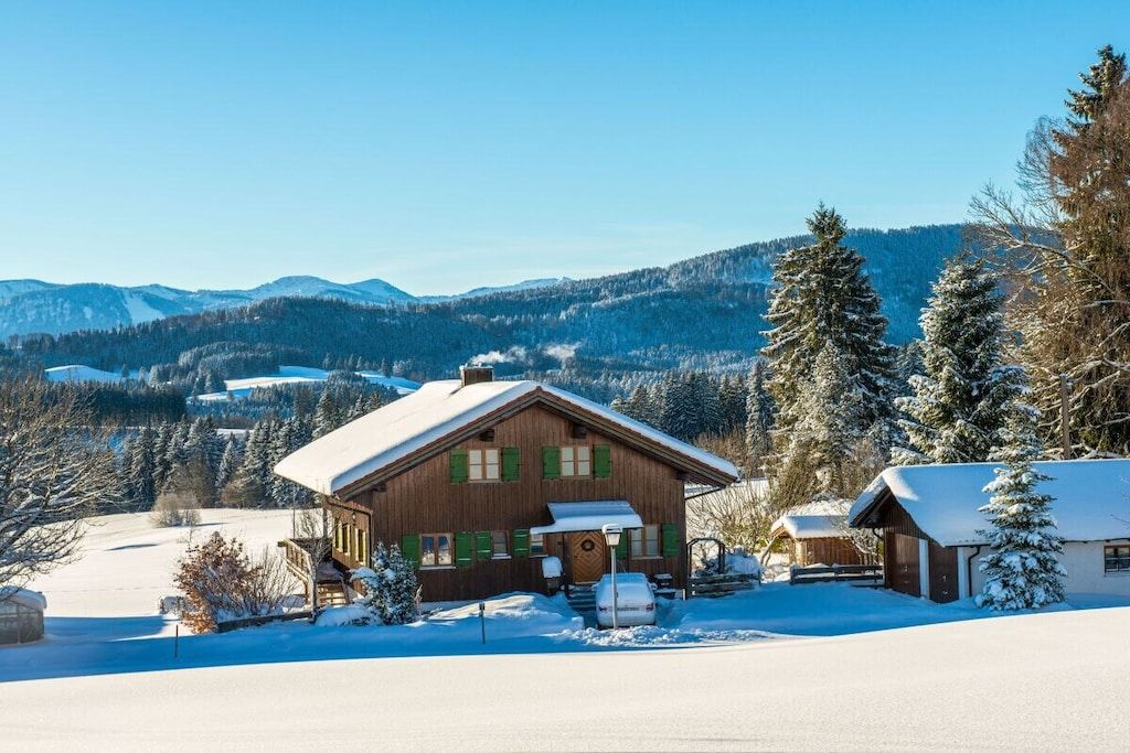 A wooden cabin with a snow-covered roof, surrounded by a snowy landscape and mountains in the background.