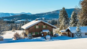 A wooden cabin with a snow-covered roof, surrounded by a snowy landscape and mountains in the background. - Holiday home with 3 bedrooms (Weitnau-Hellengerst)