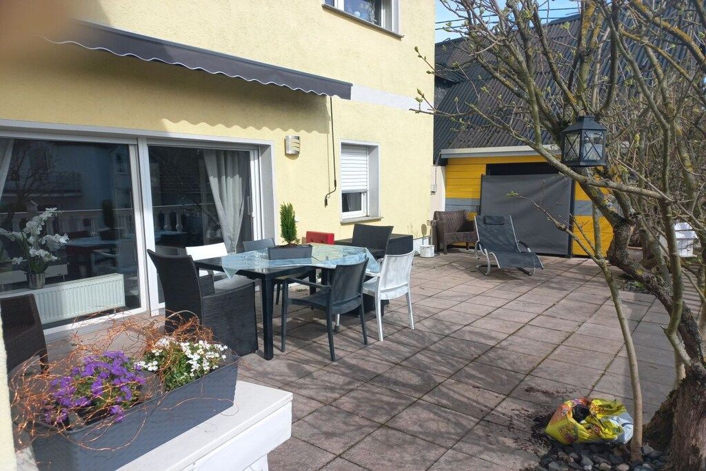 An outdoor patio with a dining table and chairs, a small garden area with flowers, and a building with a yellow facade.