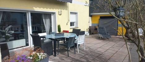 An outdoor patio with a dining table and chairs, a small garden area with flowers, and a building with a yellow facade.