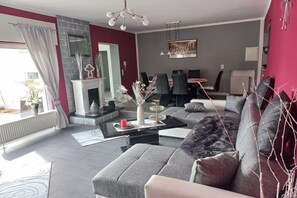 A modern living room with a fireplace, grey sofas, a dining area, and a view of the outdoors. - Holiday apartment with 3 bedrooms (Bacharach- Henschhausen)