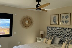 A bedroom with a bed, a ceiling fan, a wall-mounted mirror, a window with a view, and framed botanical prints. - Apartment Tortuguero (Morro Jable, Jandia, Fuerteventura)