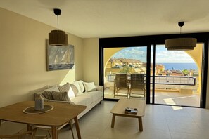 Un salon comprenant un canapé, une table basse et une table à manger. Un tableau orne le mur, et de grandes portes vitrées offrent une vue sur la mer.