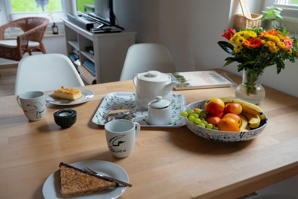 Ein Esstisch mit einem Teekessel, Tassen, einem Teller mit Kuchenstück, einer Obstschale und einer Vase mit Blumen.