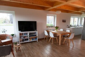 Uma sala de estar moderna com televisão de tela plana, estante, área de jantar com mesa e cadeiras de madeira, e cozinha com pia e armários.