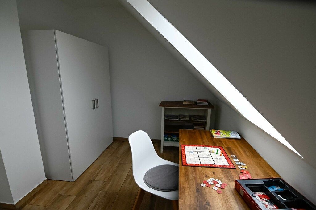 A room with a sloped ceiling, a white chair, a wooden table with a board game, and a bookshelf.