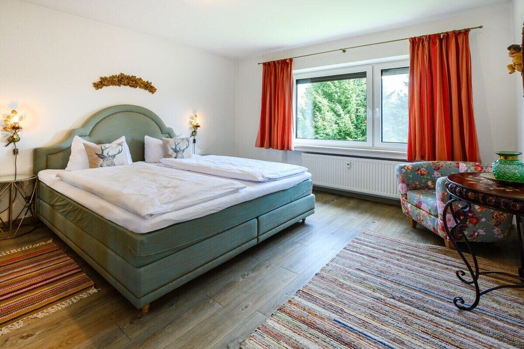 Ein Schlafzimmer mit einem Doppelbett, einem Fenster mit Vorhängen, einem Heizkörper, einem blumigen Sessel und einem kleinen Tisch mit einer Vase.