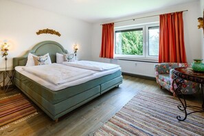 Ein Schlafzimmer mit einem Doppelbett, einem Fenster mit Vorhängen, einem Heizkörper, einem blumigen Sessel und einem kleinen Tisch mit einer Vase.