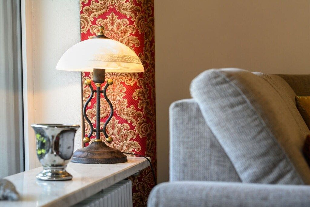 Een lamp op een tafel, een behang met patroon en een grijze bank.