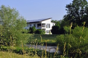 Une maison moderne à deux étages, dotée d’un balcon, entourée de verdure et traversée par un petit ruisseau.