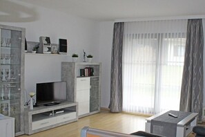 Ein modernes Wohnzimmer mit Flachbildfernseher, integrierten Regalen, einem Couchtisch und einem großen Fenster mit leichten Vorhängen.