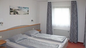 Quarto de hotel com duas camas, janela com cortinas, radiador e quadro na parede.