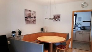 Uma área de refeições compacta com uma mesa redonda, duas cadeiras e uma pequena mesa lateral com uma planta. Um relógio é visível na parede, e uma cozinha pode ser vista através de uma porta aberta.