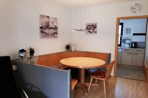 A compact dining area with a round table, two chairs, and a small side table with a plant. A clock is visible on the wall, and a kitchen area is seen through an open doorway. - Holiday home Aggenstein (Lechbruck)