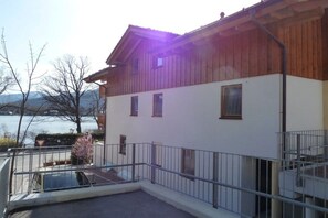 Un bâtiment à deux étages, avec un étage supérieur en bois et un étage inférieur peint en blanc, un balcon doté d’une rambarde en métal, et une vue sur une étendue d’eau et des montagnes en arrière-plan.