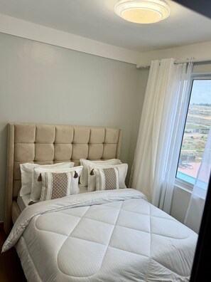 Ein Schlafzimmer mit einer gesteppten Kopfstütze, einem großen Bett mit weißer Bettwäsche und Blick auf eine Landschaft durch ein Fenster mit weißen Vorhängen.