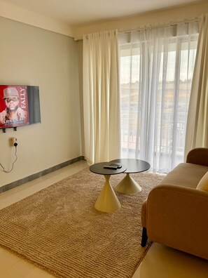 A living room with a sofa, a small round table, a TV mounted on the wall, and a window with sheer curtains. - Charming apartment in Pridelands with access to fitness room (Pridelands)