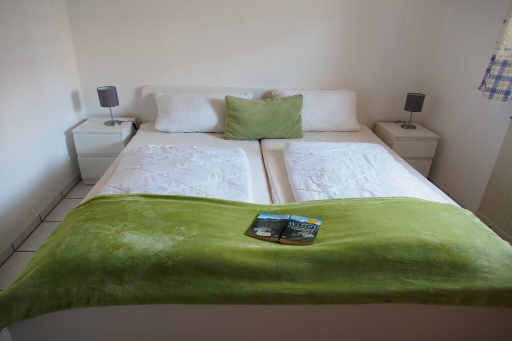 Ein Bett mit weißer Bettwäsche, einer grünen Tagesdecke und einem Buch darauf. Es gibt zwei Nachttische mit Lampen.