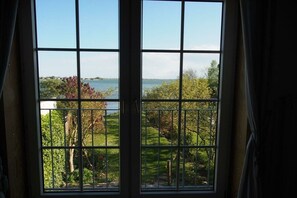 A view through a window with a metal grid, overlooking a body of water and a landscaped garden. - Holiday home in Fehmarn with swimming pool (Fehmarn)