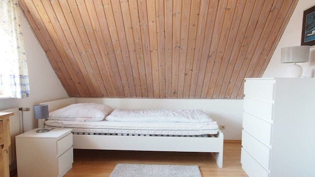 A bedroom with a sloped ceiling, a bed, a nightstand, a dresser, and a window with curtains. - Holiday home in Fehmarn with swimming pool (Fehmarn)