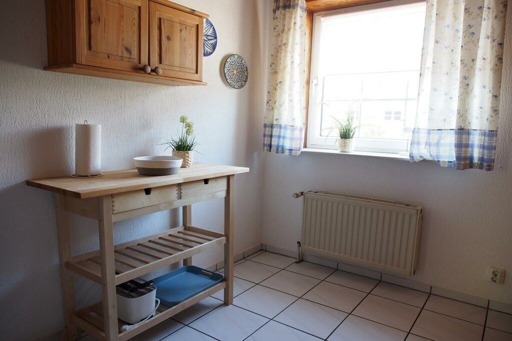 A kitchen with wooden cabinets, a white radiator, a window with floral curtains, and a wooden table with shelves.