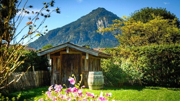 A rustic wooden cabin with a mountain backdrop, surrounded by lush greenery and flowering plants. - Holiday apartment with 2 bedrooms (Farchant)