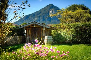 Een rustieke houten hut met een berg op de achtergrond, omgeven door weelderige begroeiing en bloeiende planten.