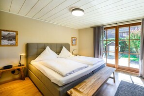 Ein Schlafzimmer mit einem Bett, Nachttischen, einer Bank, einem Fenster mit Vorhängen und Blick ins Grüne.