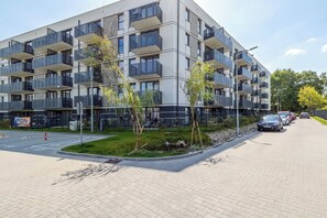 A modern multi-story residential building with balconies, a landscaped garden area, and a parking lot with cars. - Comfortable Apartment with a Balcony | Air Conditioning (Gdańsk)