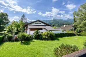A two-story house with a sloped roof, surrounded by a well-maintained garden and a view of mountains in the background. - Holiday apartment with 1 bedroom (Garmisch-Partenkirchen)