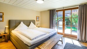 A double bed with a bench, a bedside table, and a view of a garden through a sliding door. - Holiday apartment with 1 bedroom (Farchant)