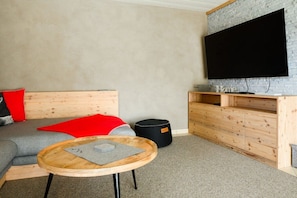 A modern living room with a grey sofa, a red throw pillow, a wooden coffee table, a black ottoman, and a mounted flat-screen TV. - Holiday apartment with 1 bedroom (Farchant)