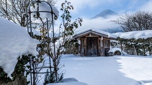 Una cabaña de madera con techo nevado, rodeada de nieve y árboles, con una montaña al fondo.