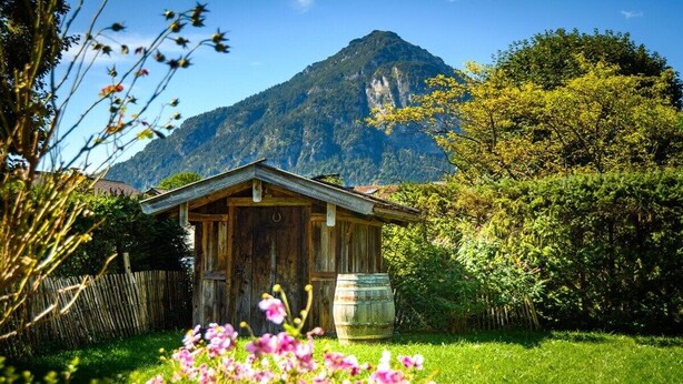 A rustic wooden cabin with a barrel in the foreground, surrounded by lush greenery and a mountain in the background. - Holiday apartment with 1 bedroom (Farchant)