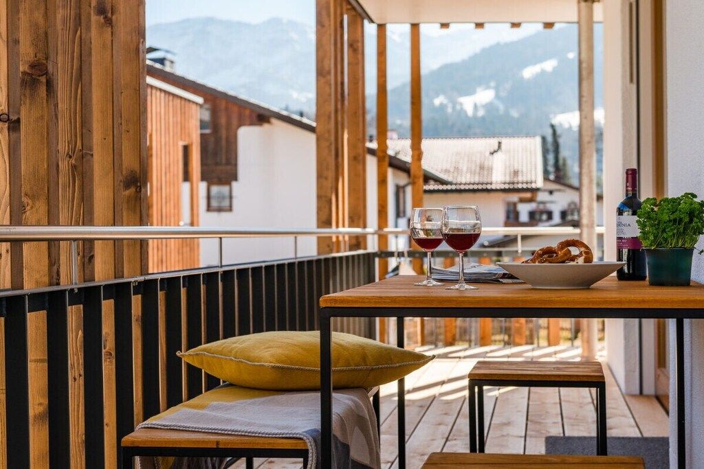Un balcón de madera con una mesa, dos copas de vino tinto, un cuenco de palitos de pretzel y una botella de vino.
