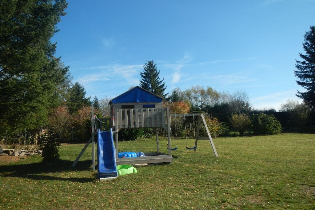 Un terrain de jeux avec des balançoires et un toboggan, entouré d’arbres et d’herbe.