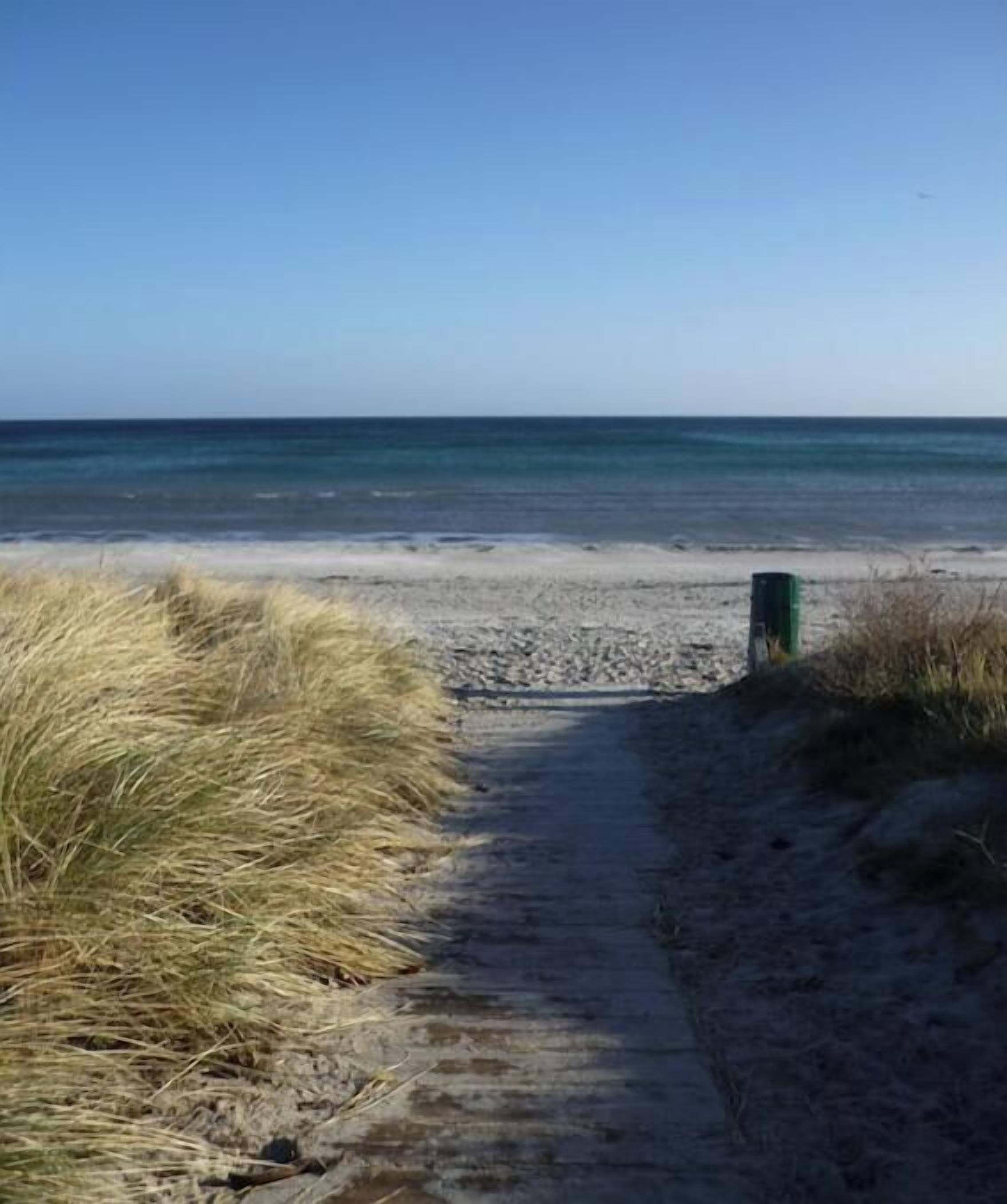 Ein sandiger Küstenpfad führt zum Meer, in der Ferne ist ein Mülleimer zu sehen.
