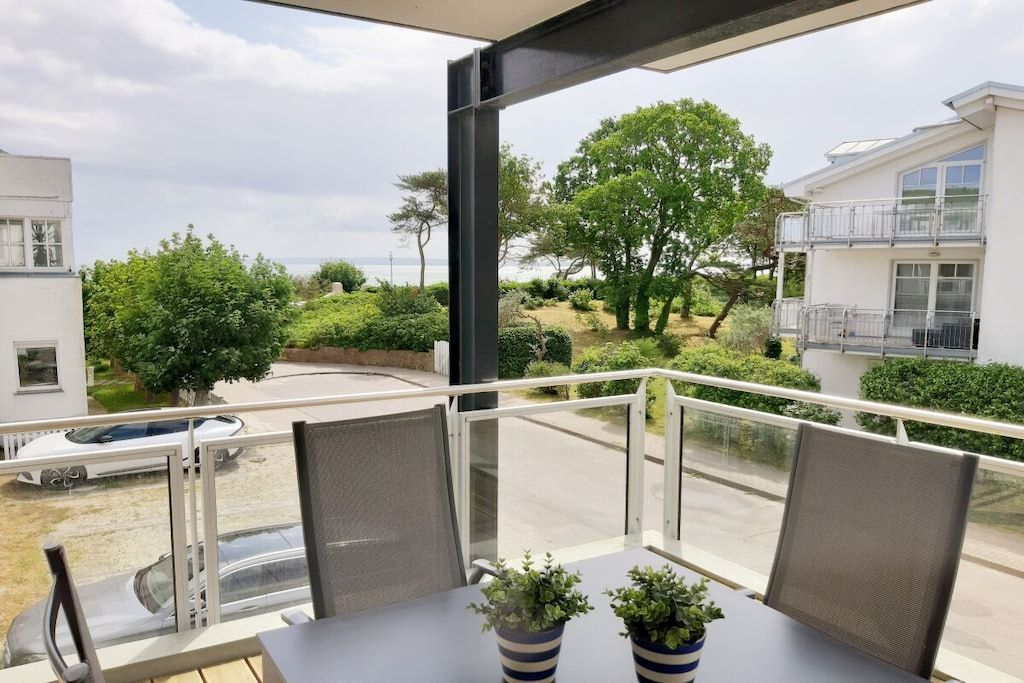 Een balkon met een tafel en stoelen, met uitzicht op een aangelegde tuin met bomen en de zee.