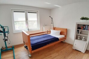 Une chambre avec un lit simple, un cadre en bois, un matelas bleu, un oreiller blanc, une lampe sur pied, une bibliothèque et une fenêtre avec des stores.