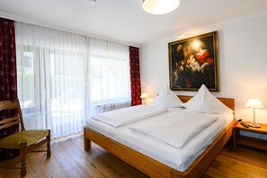A hotel room with a wooden bed, a chair, a painting on the wall, and a window with curtains. - Holiday apartment with 2 bedrooms (Garmisch-Partenkirchen)